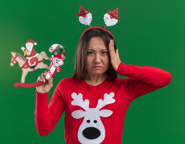 Foto gratuita joven asiática molesta con aro de pelo de navidad con suéter sosteniendo un juguete de navidad con dulces poniendo la mano en la cabeza aislada sobre fondo verde