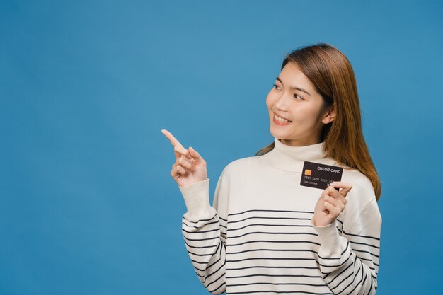 Joven asiática muestra tarjeta bancaria de crédito con expresión positiva, sonríe ampliamente, vestida con ropa informal sintiendo felicidad y parada aislada en la pared azul