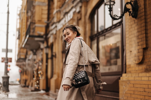 Joven asiática de muy buen humor camina por la ciudad en elegante gabardina con una pequeña bolsa negra