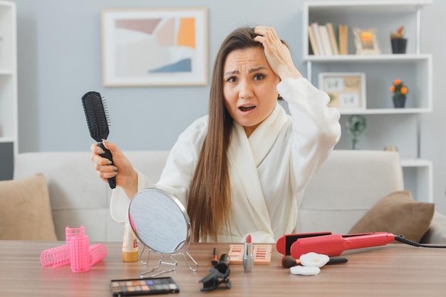 Joven asiática con el pelo largo y oscuro sentada en el tocador de su casa haciendo la rutina de maquillaje matutino luciendo confundida mientras se cepilla el pelo sufriendo de pérdida de cabello