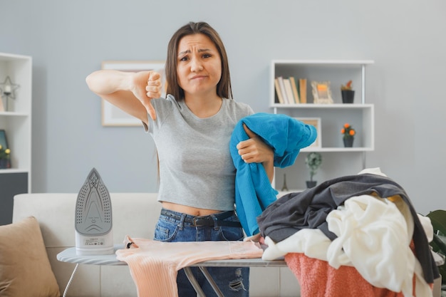 Foto gratuita joven asiática con una pila de ropa planchada con plancha eléctrica en la tabla de planchar en el interior de su casa preocupada mostrando el pulgar hacia abajo