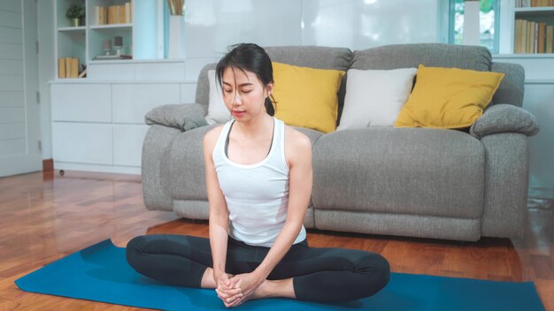 Joven asiática practicando yoga en la sala de estar. Hembra hermosa atractiva que se resuelve para sano en casa. Concepto de ejercicio de estilo de vida mujer.