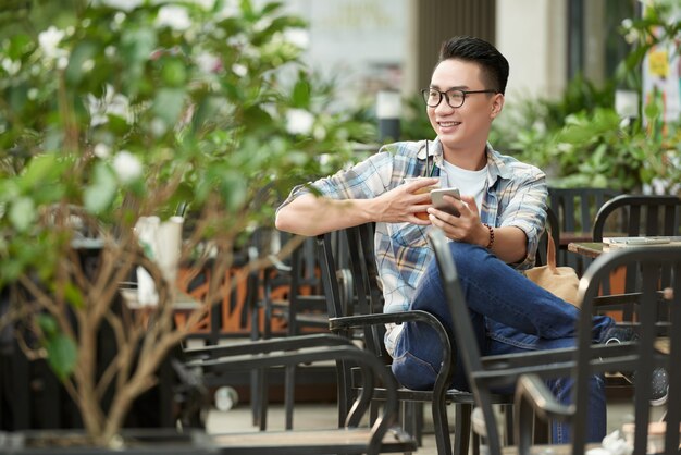 Joven asiática relajante en el café al aire libre con teléfono inteligente y té