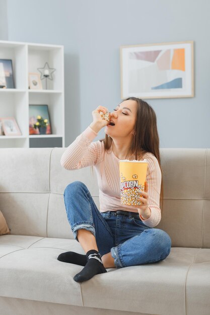 Joven asiática con ropa informal sentada en un sofá en el interior de su casa con un cubo de palomitas de maíz feliz y positiva relajándose descansando