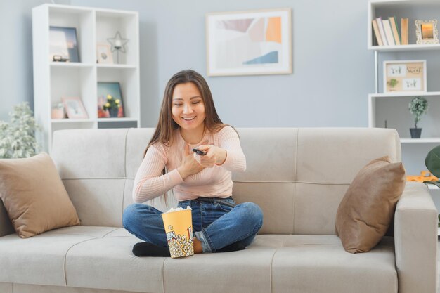 Joven asiática con ropa informal sentada en un sofá en el interior de su casa con un cubo de palomitas de maíz sosteniendo un control remoto viendo la televisión feliz y positiva sonriendo alegremente