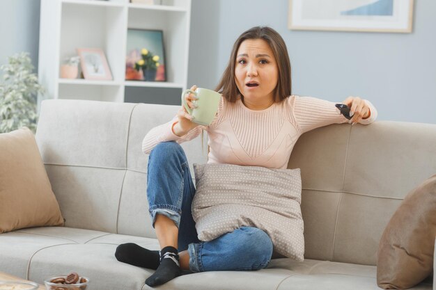 joven asiática con ropa informal sentada en un sofá en el interior de su casa sosteniendo un control remoto viendo la televisión bebiendo té de una taza sorprendida