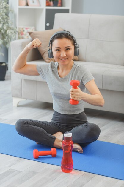 Joven asiática saludable con auriculares haciendo ejercicio en casa con una botella de agua y pesas en la sala de estar sentada en una alfombra de yoga mostrando su fuerza y bíceps