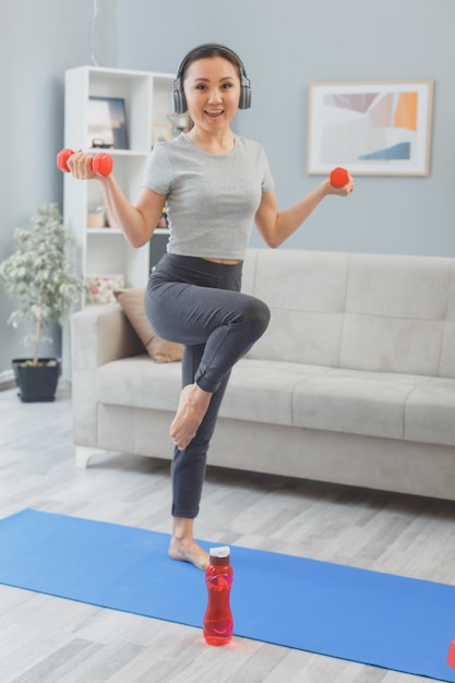 Joven asiática sana con auriculares haciendo ejercicio en casa con una botella de agua y pesas levantando la pierna en una alfombra de yoga feliz y positiva sonriendo alegremente