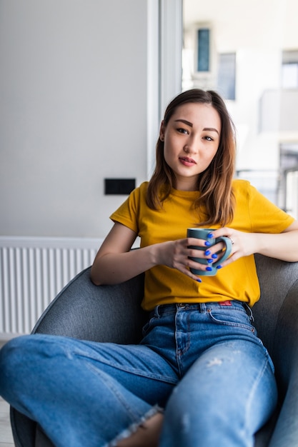 Joven asiática sentada en una silla moderna, disfrutando de un café delante de la ventana