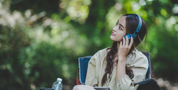 Una joven asiática se sienta en una silla cerca del arroyo escuchando música con auriculares inalámbricos y usa una tableta felizmente mientras acampa en el espacio de copia del bosque