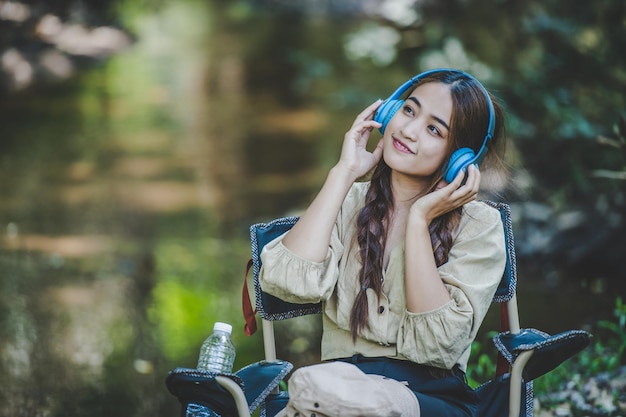 Una joven asiática se sienta en una silla cerca del arroyo escuchando música con auriculares inalámbricos y usa una tableta felizmente mientras acampa en el espacio de copia del bosque