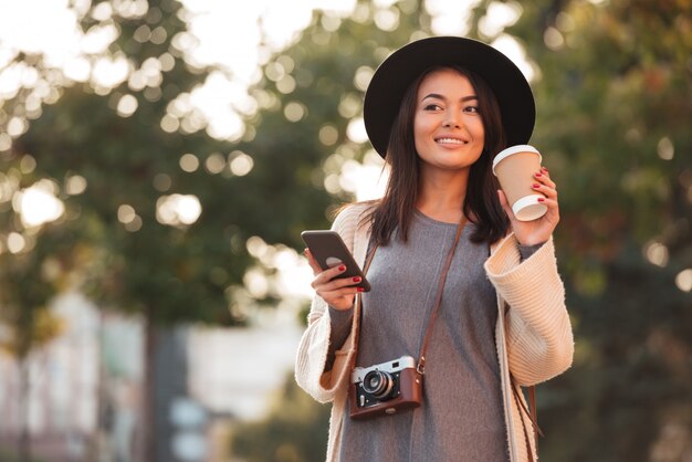 Joven asiática en sombrero negro tomando café y sosteniendo el teléfono móvil mientras camina en el parque al aire libre