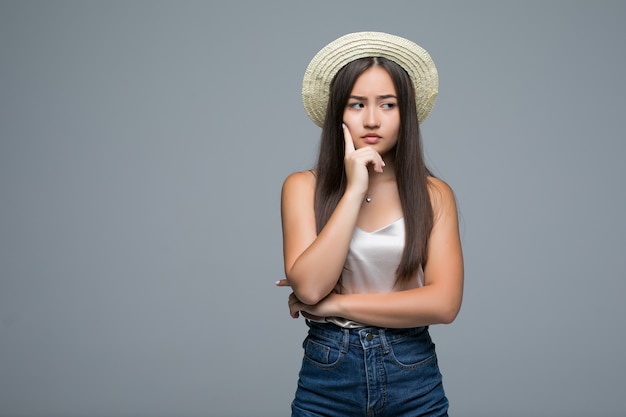 Joven asiática en sombrero de paja pensando en algo sobre fondo gris