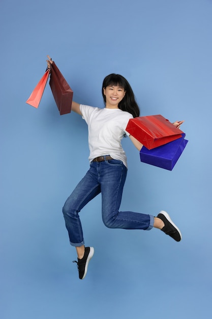 Joven asiática sosteniendo bolsas de la compra y saltando sobre azul