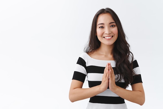 Una joven asiática tierna, alegre y amistosa, pacífica, se toma de la mano para orar y sonríe, agradecida, expresa gratitud, agradeciendo la ayuda, de pie, fondo blanco, aprecia el esfuerzo