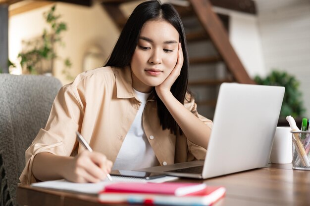 Joven asiática tomando notas escribiendo y haciendo la tarea sentado cerca de la computadora portátil mujer coreana trabajando fr ...