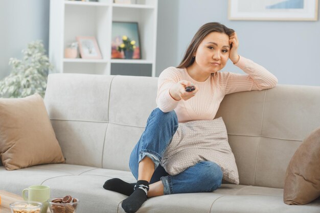 joven asiática vestida de forma informal sentada en un sofá en el interior de su casa sosteniendo un televisor remoto con expresión escéptica