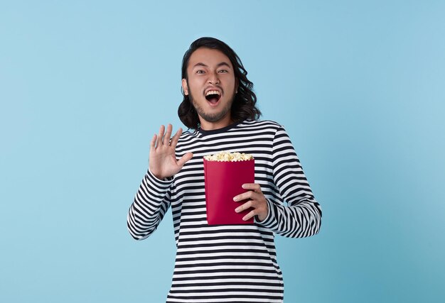 Foto gratuita un joven asiático fresco ve una película y sostiene un cubo de palomitas de maíz aisladas sobre fondo azul