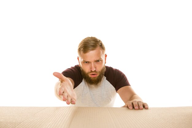 Joven asombrado abriendo el paquete postal más grande aislado en blanco. Modelo masculino sorprendido en la parte superior de la caja de cartón mirando hacia adentro.