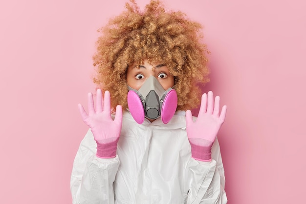 Una joven asustada e impresionada mantiene las palmas levantadas hacia la cámara y trata de protegerse del peligro vestida con guantes protectores de traje químico blanco y máscara de gas aislada sobre fondo rosa