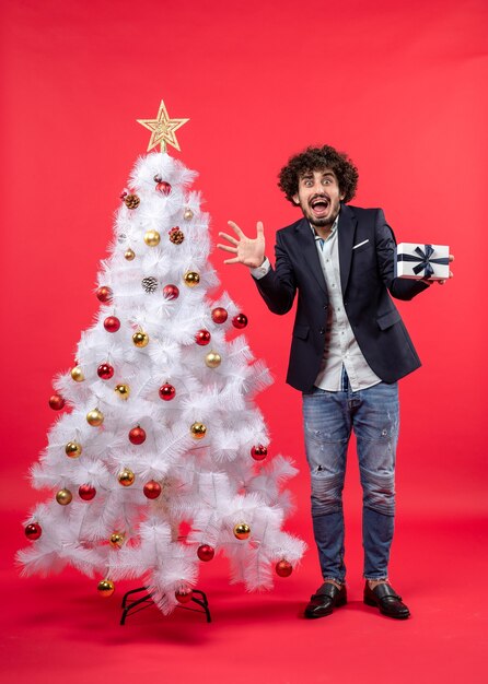 Joven asustado sosteniendo su regalo de pie cerca del árbol de Navidad blanco decorado en el lado derecho de rojo