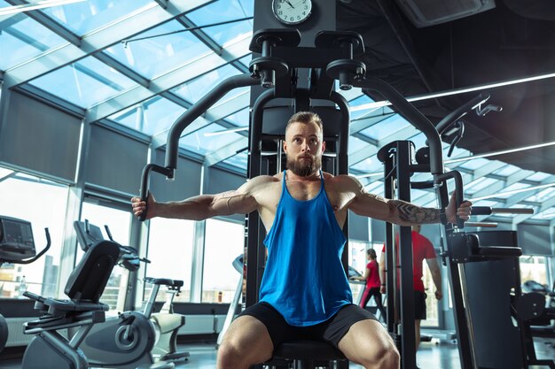 Joven atleta caucásico muscular entrenando en el gimnasio, haciendo ejercicios de fuerza, practicando, trabajando en la parte superior de su cuerpo, tirando de pesas y pesas. Fitness, bienestar, concepto de estilo de vida saludable, trabajo.