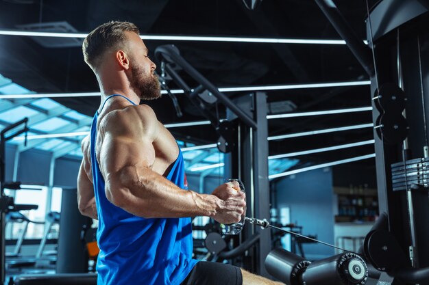 Joven atleta caucásico muscular entrenando en el gimnasio, haciendo ejercicios de fuerza, practicando, trabajando en la parte superior de su cuerpo, tirando de pesas y pesas. Fitness, bienestar, concepto de estilo de vida saludable, trabajo.