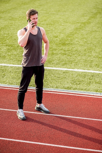 Joven atleta masculino de pie en la pista de carreras hablando por teléfono móvil