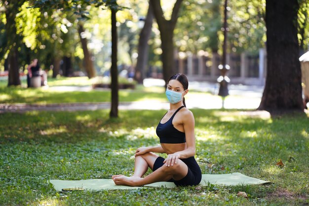 Joven atlética con una máscara protectora médica, haciendo yoga en el parque por la mañana, entrenamiento de la mujer en una estera de yoga