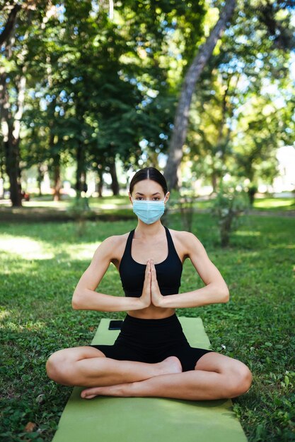 Joven atlética con una máscara protectora médica, haciendo yoga en el parque por la mañana