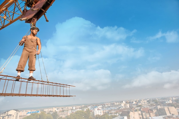 Joven atlético en ropa de trabajo y sombrero de pie en la construcción en lo alto y mirando a otro lado. Paisaje urbano y cielo azul de fondo. Grúa de construcción grande que sostiene la construcción con el hombre sobre la ciudad en el aire.