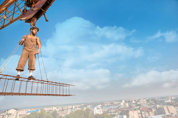 Joven atlético en ropa de trabajo y sombrero de pie en la construcción en lo alto y mirando a otro lado. Paisaje urbano y cielo azul de fondo. Grúa de construcción grande que sostiene la construcción con el hombre sobre la ciudad en el aire.