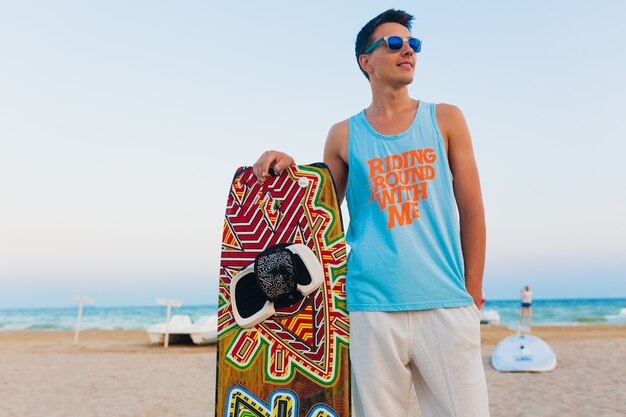 Joven atlético con tabla de kitesurf posando en la playa con gafas de sol en vacaciones de verano