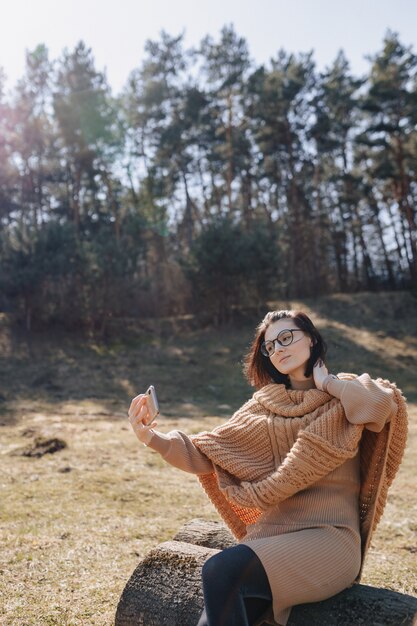 Joven atractiva chica elegante en la naturaleza en la pared del bosque con un teléfono en un día soleado tomando fotos de sí misma. vacaciones al aire libre y dependencia de la tecnología.