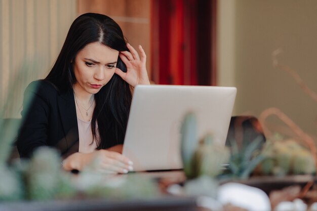 Joven atractiva chica emocional en ropa de estilo empresarial sentado en un escritorio en una computadora portátil y teléfono en la oficina o auditorio