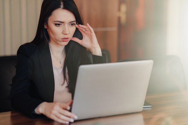 Joven atractiva chica emocional en ropa de estilo empresarial sentado en un escritorio en una computadora portátil y teléfono en la oficina o auditorio