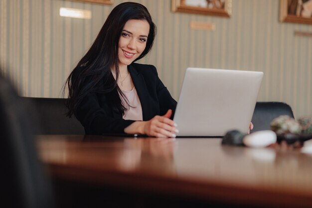 Joven atractiva chica emocional en ropa de estilo empresarial sentado en un escritorio en una computadora portátil y teléfono en la oficina o auditorio