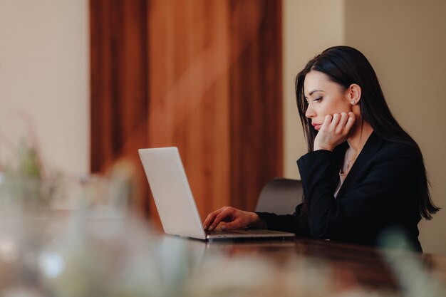 Joven atractiva chica emocional en ropa de estilo empresarial sentado en un escritorio en una computadora portátil y teléfono en la oficina o auditorio