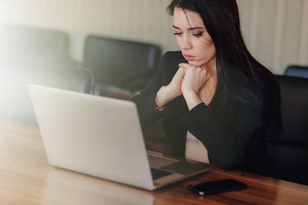 Joven atractiva chica emocional en ropa de estilo empresarial sentado en un escritorio en una computadora portátil y teléfono en la oficina o auditorio