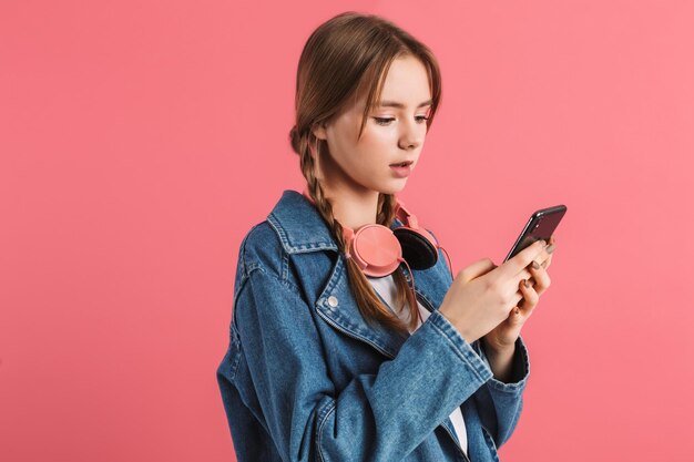 Joven atractiva chica soñadora con dos trenzas en chaqueta de mezclilla con auriculares cuidadosamente usando celular sobre fondo rosa aislado