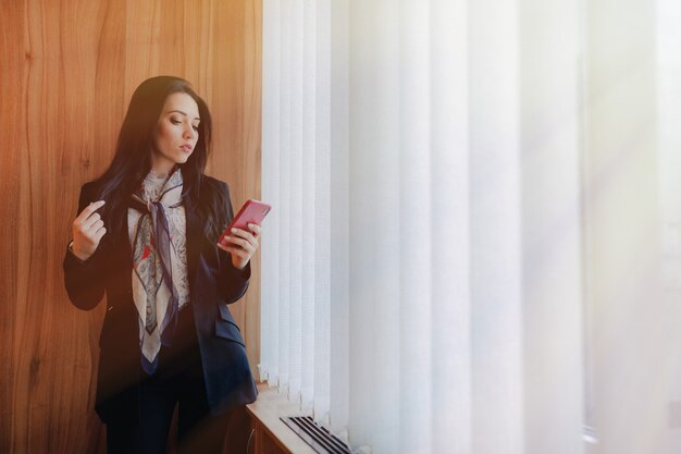 Joven atractiva emocional en ropa de estilo empresarial en una ventana con un teléfono en una oficina o auditorio moderno