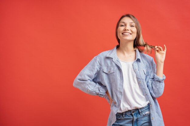 Joven atractiva jugando con su cabello y sonriendo mientras está de pie contra la pared roja