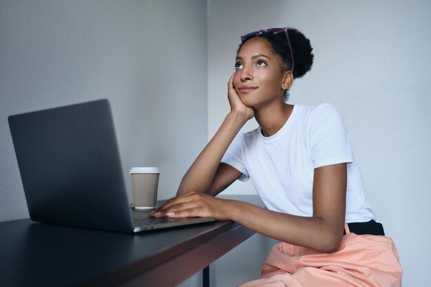 Joven y atractiva mujer afroamericana informal trabajando soñadoramente en una laptop con café en un moderno espacio de trabajo conjunto