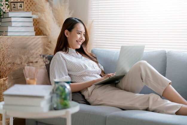 Una joven y atractiva mujer asiática que trabaja en casa usa una computadora portátil revisando el programa de búsqueda informe de planificación del proceso de trabajo con el ocio, relájese en el sofá en la sala de estar con la luz de la mañana en un momento de paz