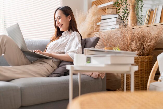 Una joven y atractiva mujer asiática que trabaja en casa usa una computadora portátil revisando el programa de búsqueda informe de planificación del proceso de trabajo con el ocio, relájese en el sofá en la sala de estar con la luz de la mañana en un momento de paz