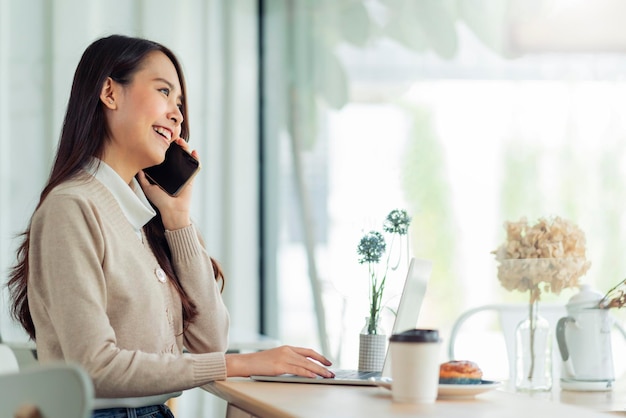 Joven y atractiva mujer asiática usa ropa casual, usa la mano, usa un teléfono inteligente y un dispositivo portátil, comunica el contacto social en el restaurante de la cafetería, siéntate en el mostrador junto a la luz del día de la ventana grande