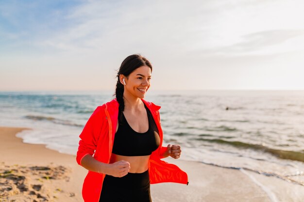 Joven y atractiva mujer delgada haciendo ejercicios deportivos en el amanecer de la mañana trotando en la playa del mar en ropa deportiva, estilo de vida saludable, escuchando música en auriculares, con chaqueta cortavientos rosa