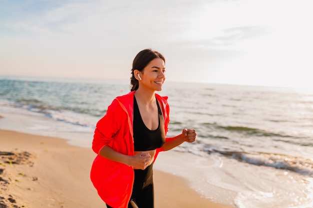 Joven y atractiva mujer delgada haciendo ejercicios deportivos en el amanecer de la mañana trotando en la playa del mar en ropa deportiva, estilo de vida saludable, escuchando música en auriculares, con chaqueta cortavientos rosa