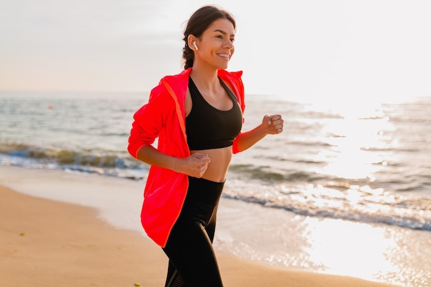 Joven y atractiva mujer delgada haciendo ejercicios deportivos en el amanecer de la mañana trotando en la playa del mar en ropa deportiva, estilo de vida saludable, escuchando música en auriculares, con chaqueta cortavientos rosa