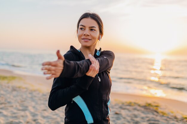 Joven y atractiva mujer delgada haciendo ejercicios deportivos en la playa del amanecer de la mañana en ropa deportiva, estilo de vida saludable, escuchando música en auriculares, haciendo estiramientos para las manos